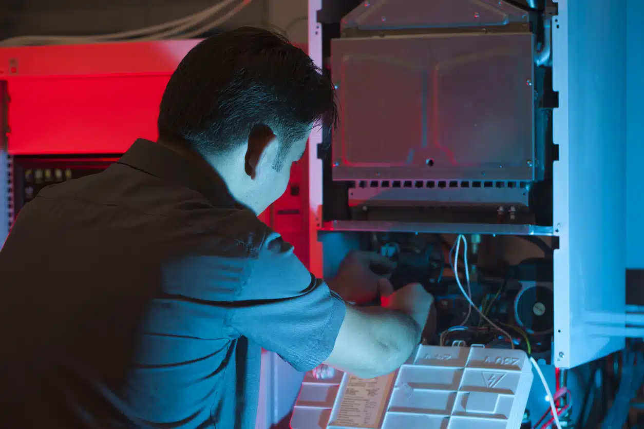 A technician works on a piece of HVAC equipment, accessing wires and components behind a removed panel.